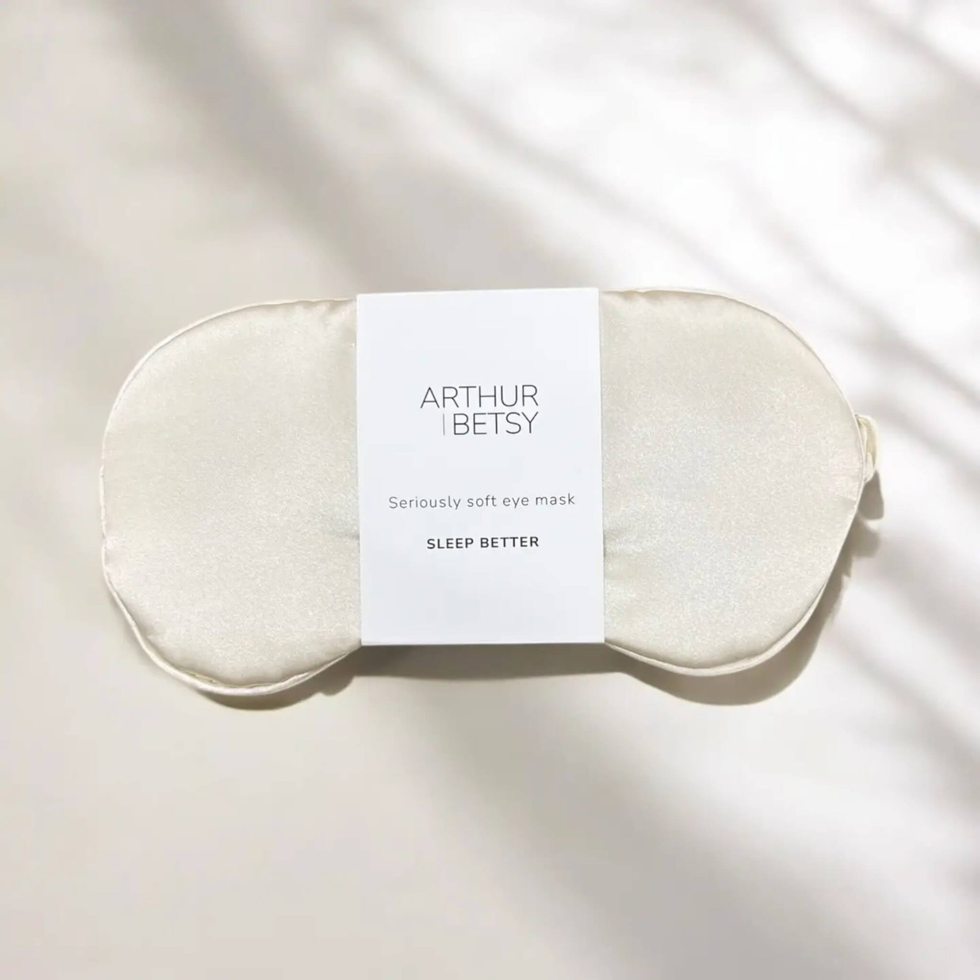 Flat lay of ivory satin sleep mask by Arthur Betsy, resting on a soft cream surface in natural light
