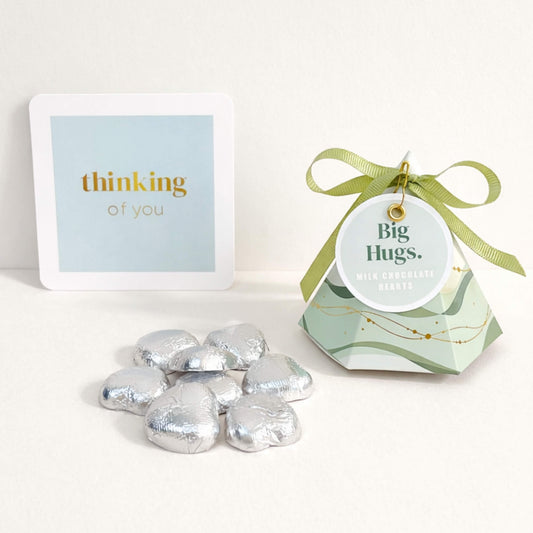 A green big hugs pyramid gift box filled with milk chocolate hearts, tied with green ribbon, with silver foil wrapped chocolate hearts arranged beside it