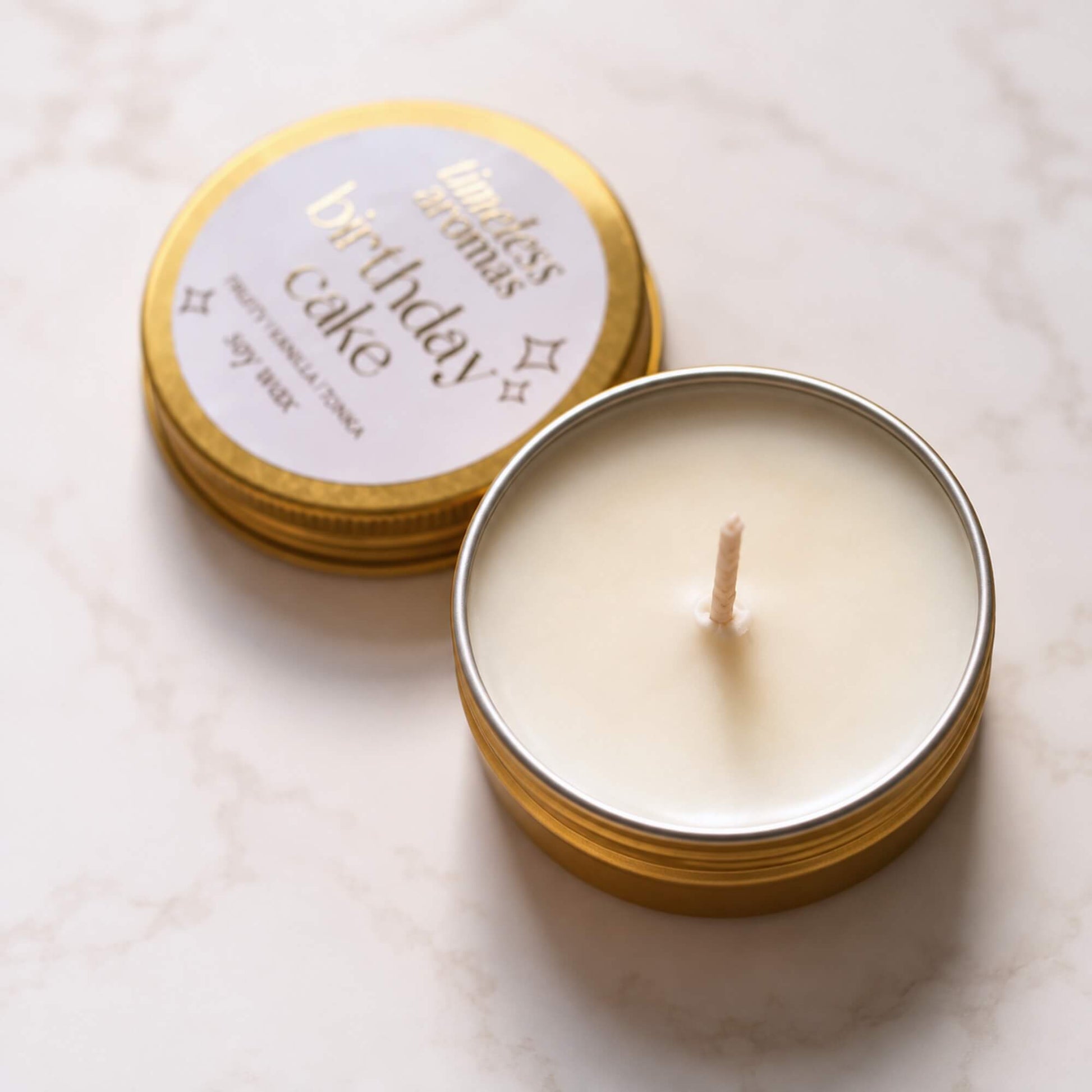 Open Birthday Cake mini soy wax candle with gold tin lid on a light marble background