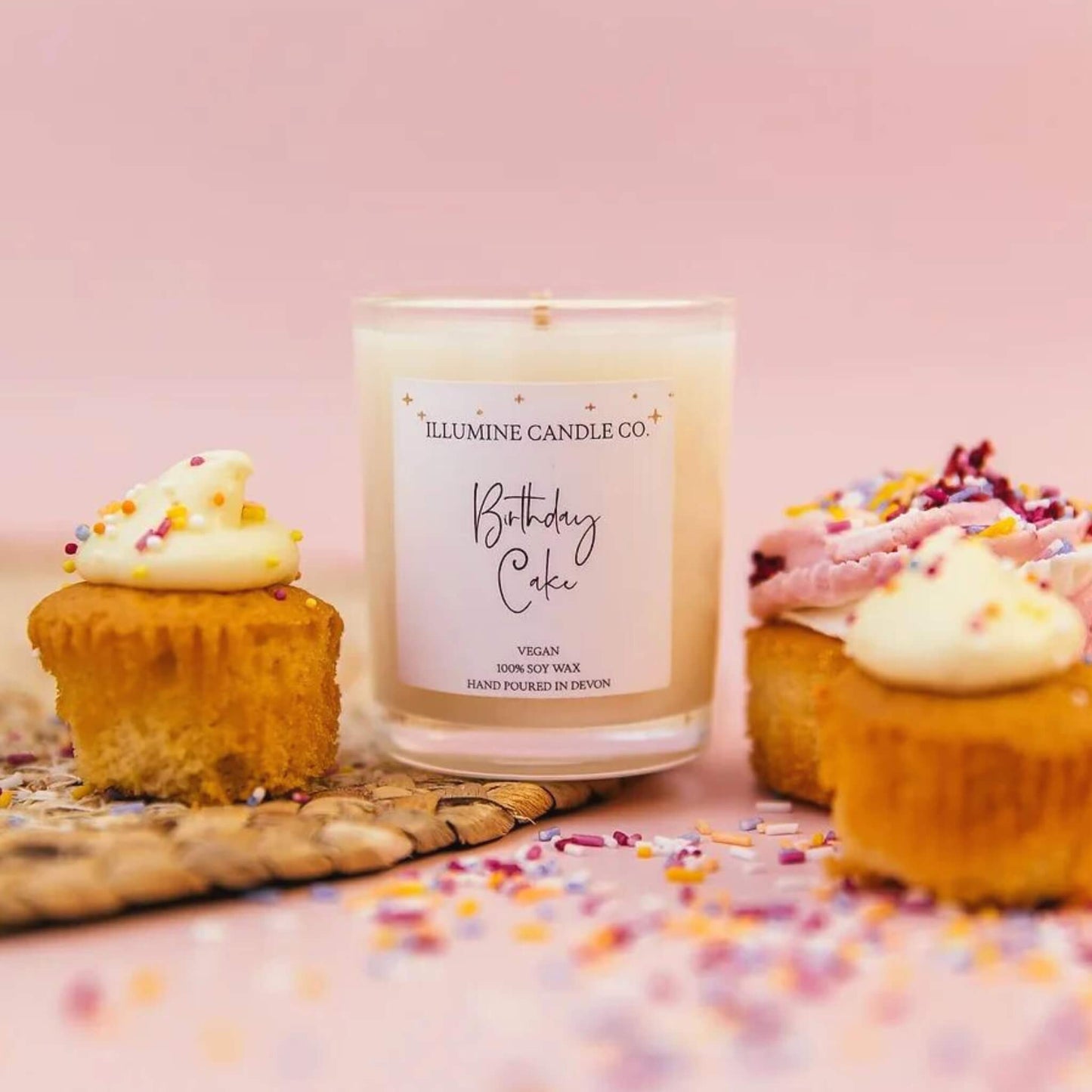 Birthday cake scented soy wax candle in a glass jar with a white label surrounded by cupcakes