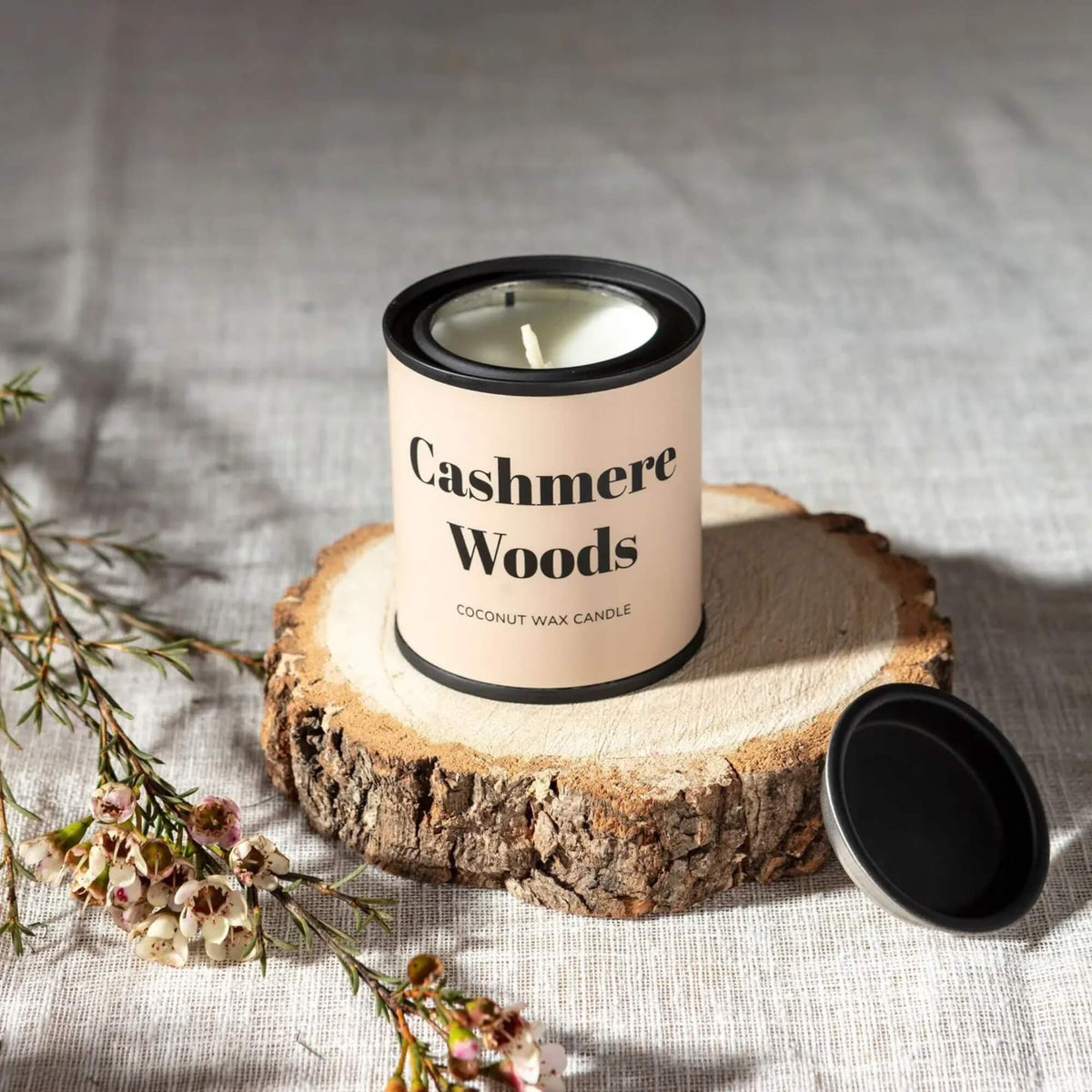 Cashmere Woods coconut wax tin candle displayed on a wooden slice with its black lid beside it