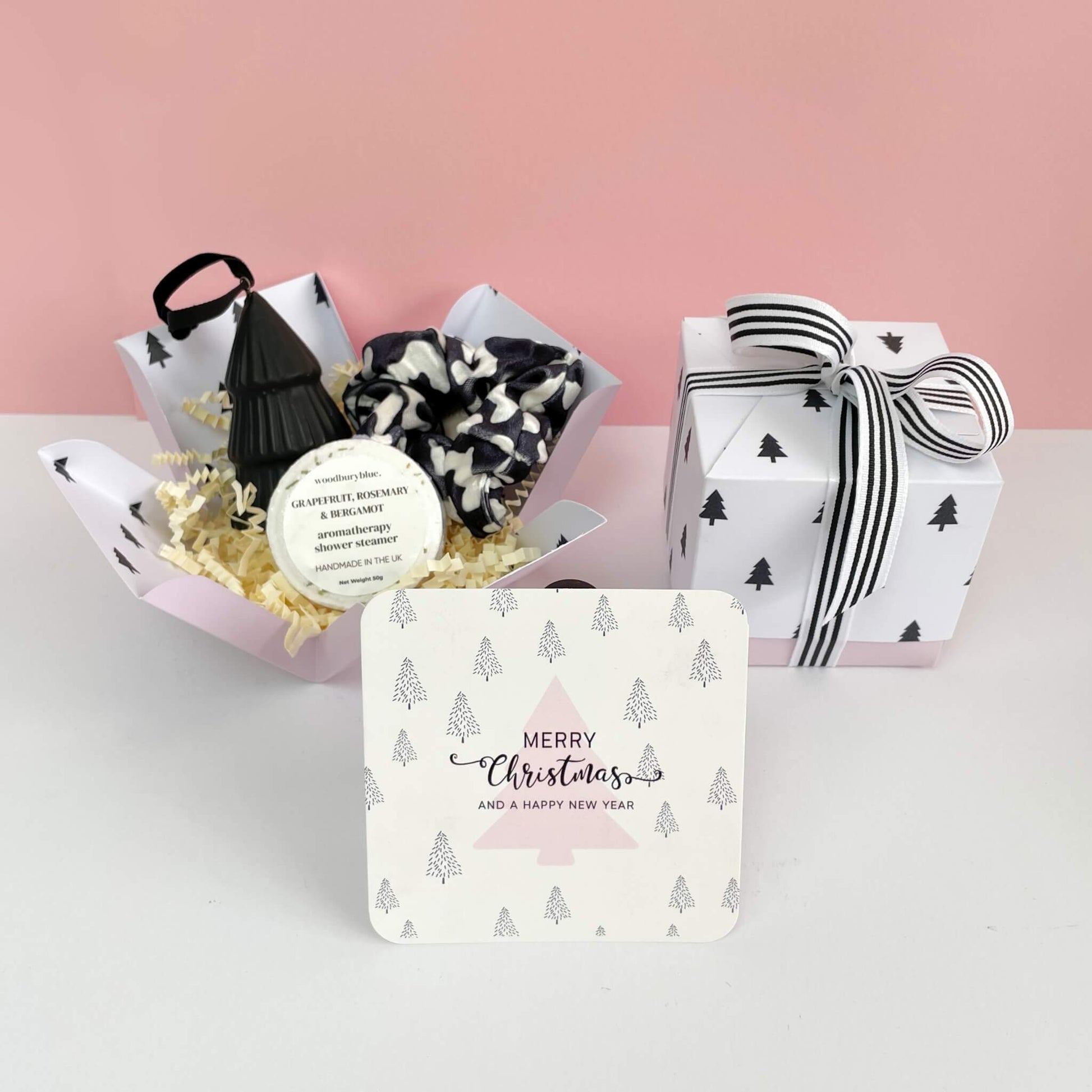 Unboxed Festive Treats Gift Box featuring a grapefruit, rosemary & bergamot aromatherapy shower steamer, black and white scrunchie, ceramic black tree ornament, and a pink tree Christmas notecard, with the ribbon-wrapped box in the background.