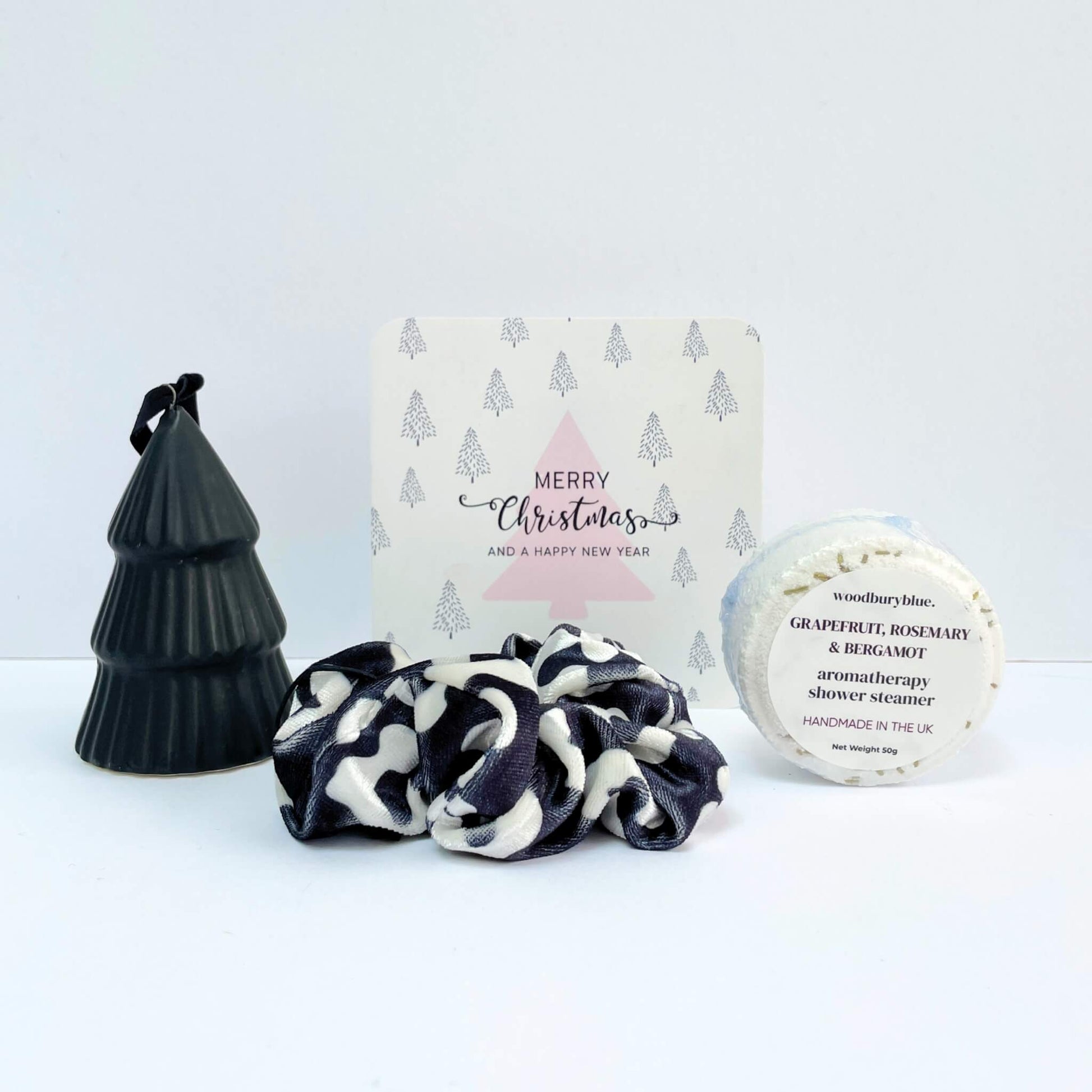 Little Box of Festive Treats featuring a grapefruit rosemary shower steamer, black and cream scrunchie, black ceramic tree ornament, and a pink tree Merry Christmas notecard.