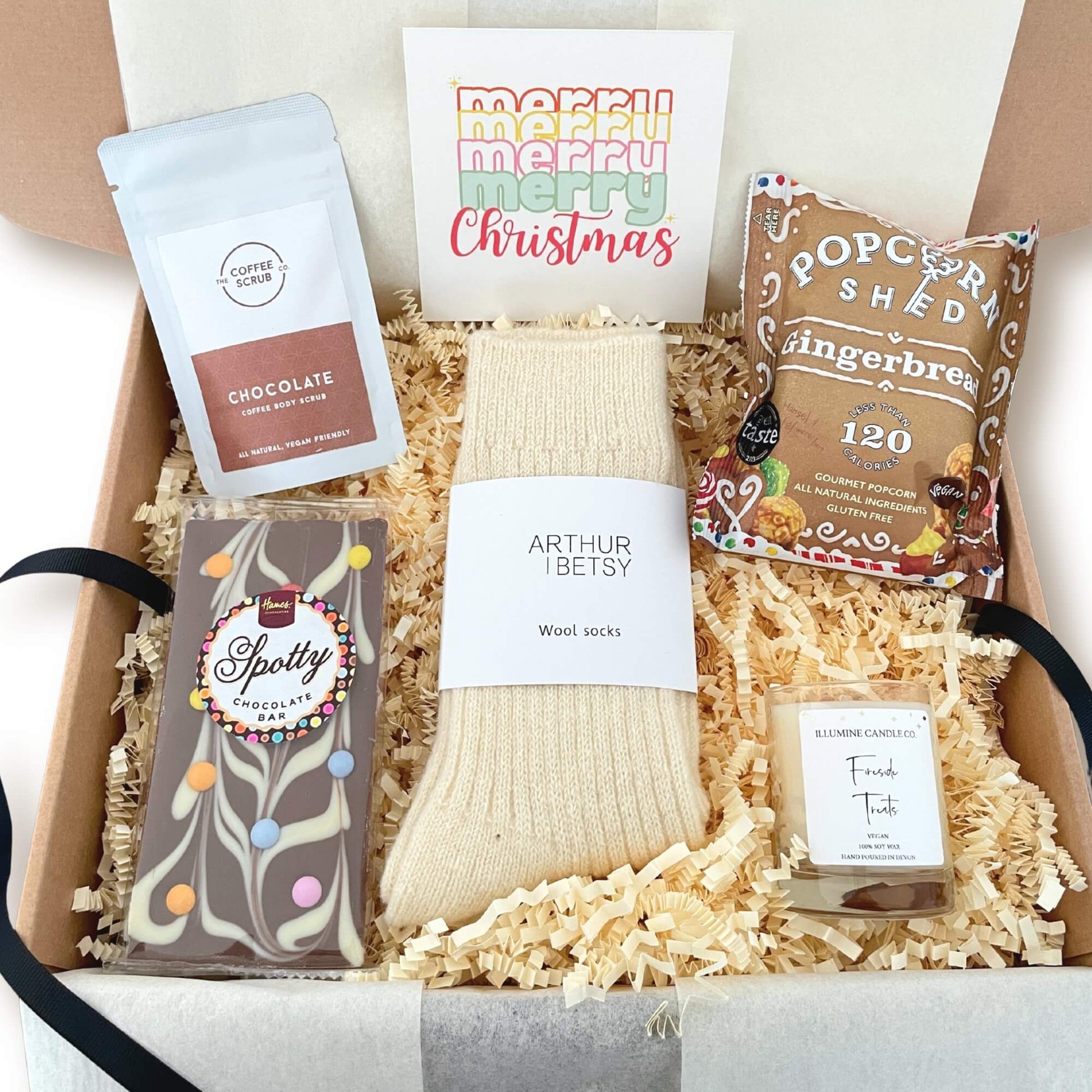Christmas gift box called Fireside Treats filled with wool socks, chocolate bar, coffee body scrub, gingerbread popcorn and a candle, presented in a kraft box with ribbon and tissue.