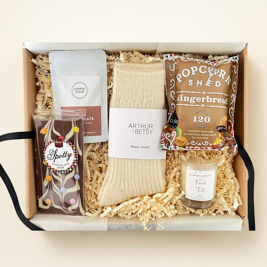 Top down photo of the Fireside Treats cosy Christmas hamper including wool socks, festive chocolate, popcorn, a scented candle and coffee scrub arranged in shredded paper.