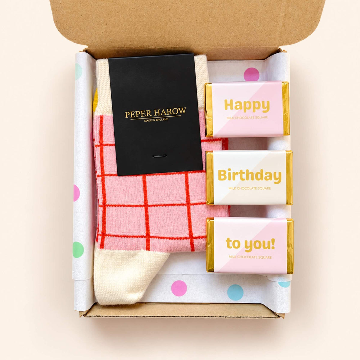 Close-up of Peper Harow pink checked cotton socks and three individually wrapped birthday milk chocolates inside a kraft letterbox gift box.