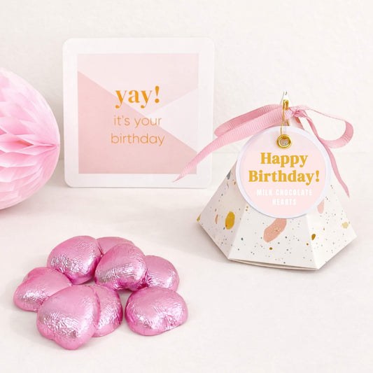 A pink happy birthday pyramid gift box containing milk chocolate hearts, tied with a pink ribbon, with pink foil wrapped chocolate hearts placed in front