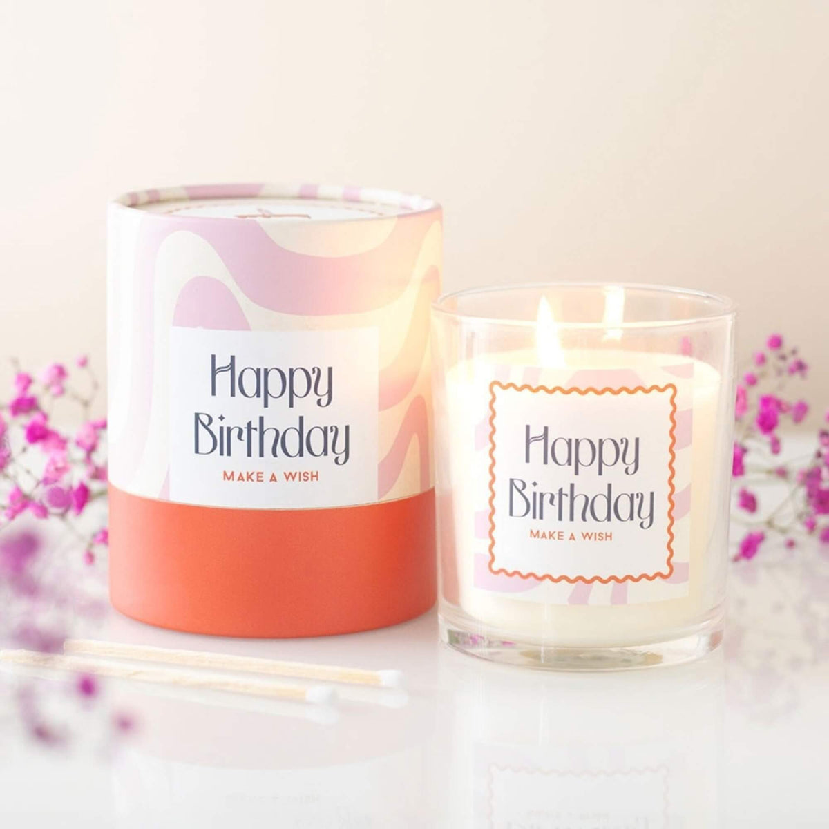 Lit Happy Birthday Vanilla Cake scented candle beside its colourful tubular gift box