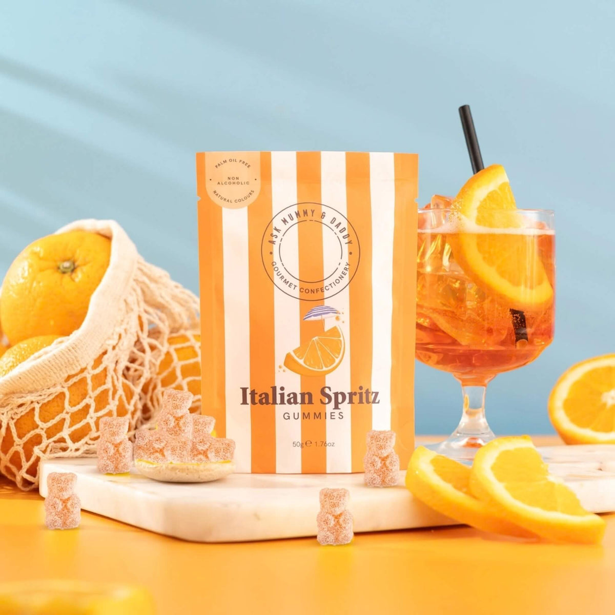 Italian Spritz gummy sweets in orange striped packaging beside an orange cocktail, fresh orange slices and gummy bears.