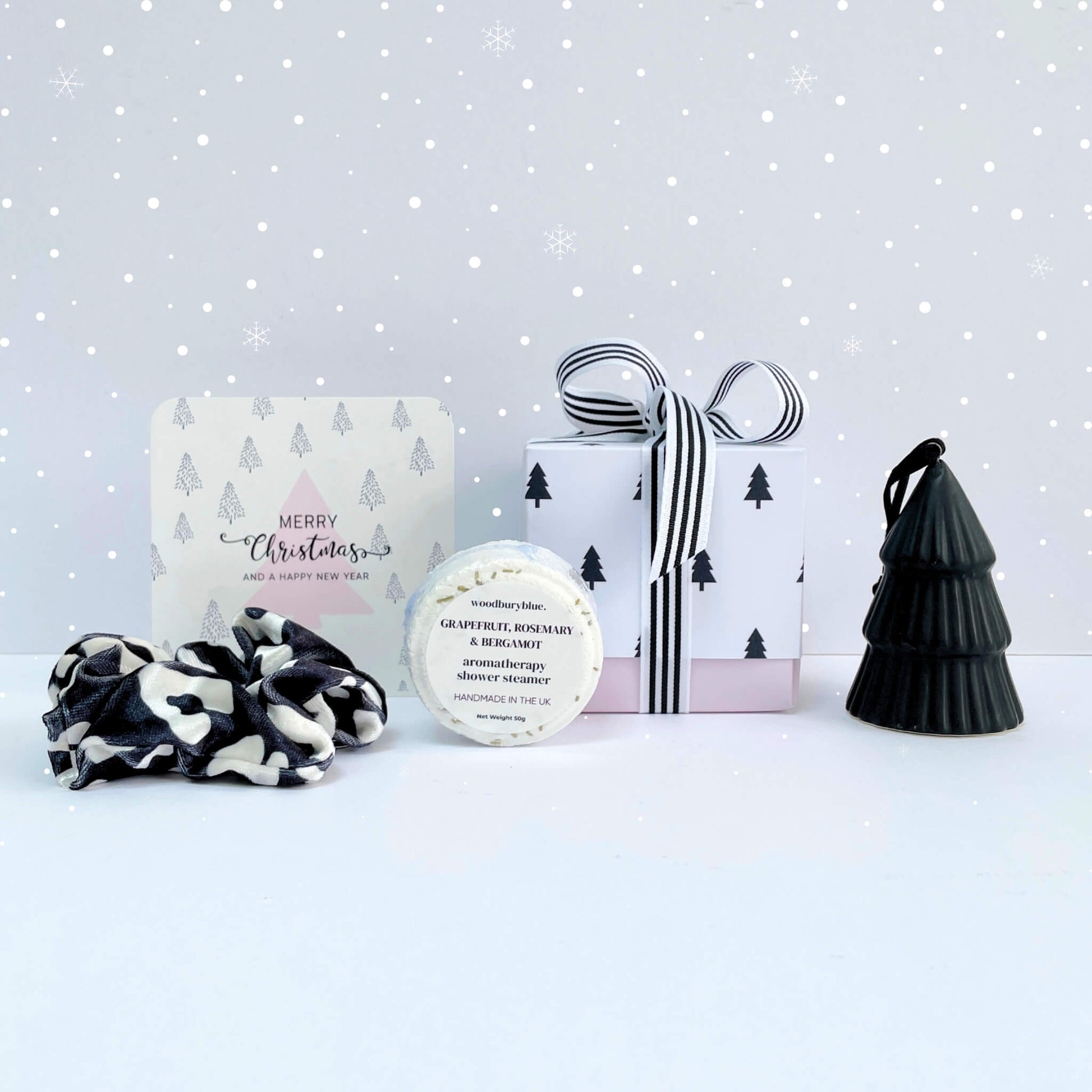Little Box of Festive Treats styled on a snowy background with striped ribbon gift box, grapefruit rosemary shower steamer, monochrome scrunchie, pink tree card, and black tree ornament.