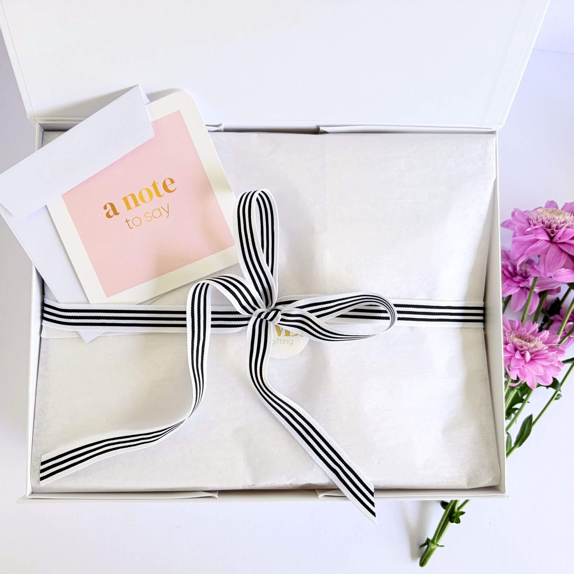 Luxury birthday gift box packaging with white tissue paper, black striped ribbon, handwritten note card and pink flowers.