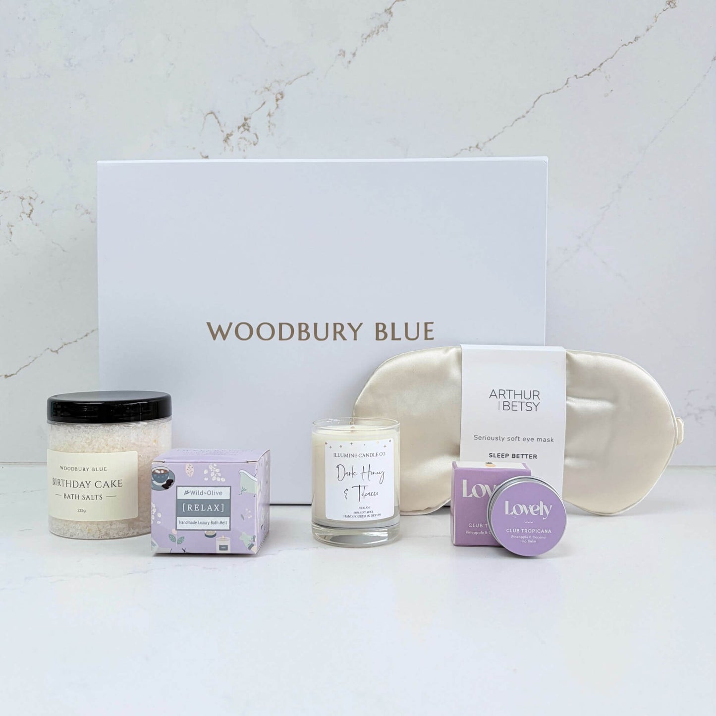 Luxury birthday pamper gift box contents with silk eye mask, birthday cake bath salts, candle, bath melt and lip balm beside a white Woodbury Blue gift box.