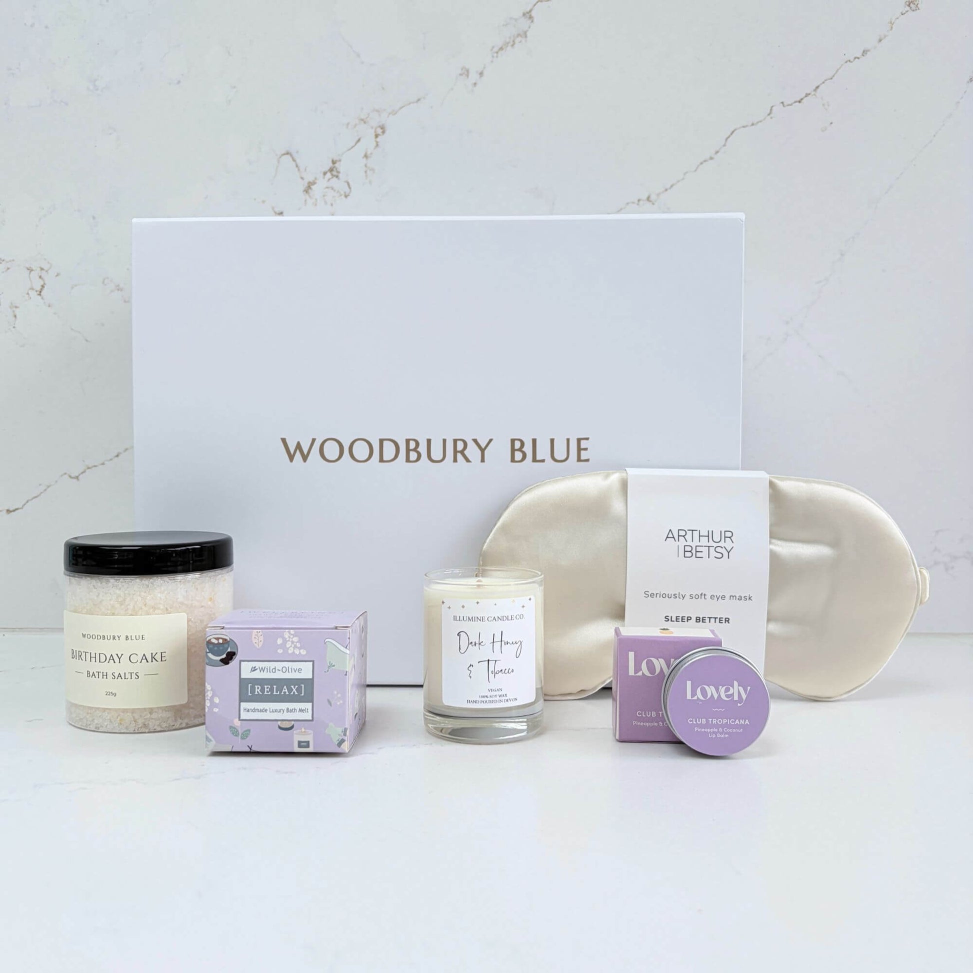 Luxury birthday pamper gift box contents with silk eye mask, birthday cake bath salts, candle, bath melt and lip balm beside a white Woodbury Blue gift box.