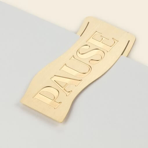 Close-up of the brushed brass Papier 'Pause' Bookmark resting on the edge of a book.