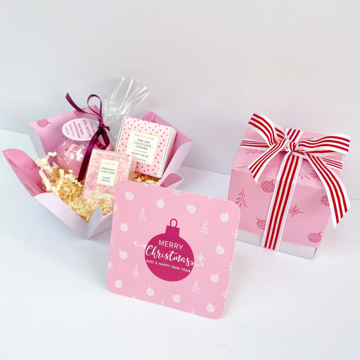 Open Rosy Glow Christmas Gift Box filled with a pink bath bomb, rosewood & ylang ylang soap, pink gin chocolate creams, and a matching bauble Christmas notecard with the ribbon-wrapped box behind.