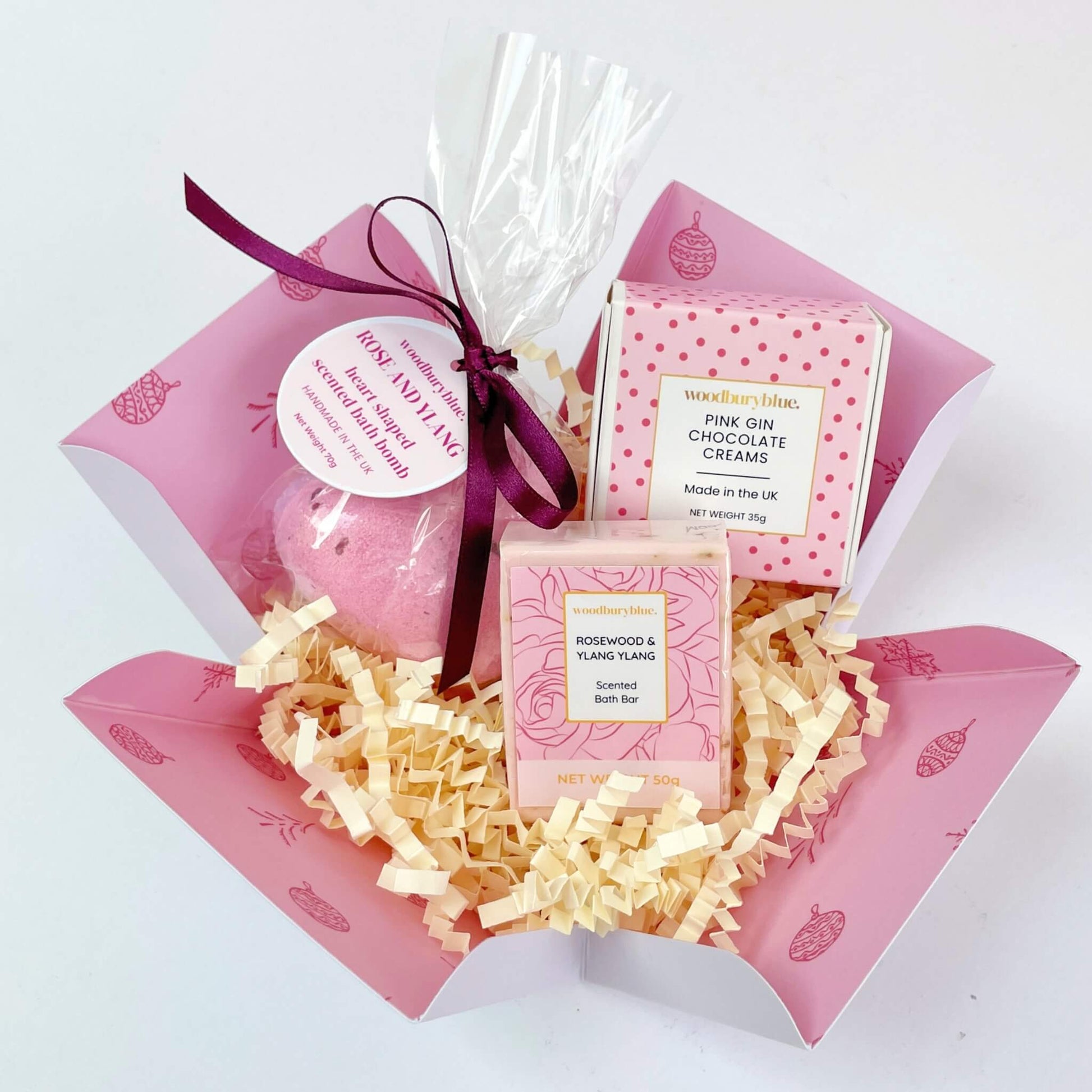 Rosy Glow Christmas Gift Box shown open with rose and ylang ylang heart-shaped bath bomb, rosewood soap bar, and pink gin chocolate creams nestled in shredded paper inside a festive pink box.