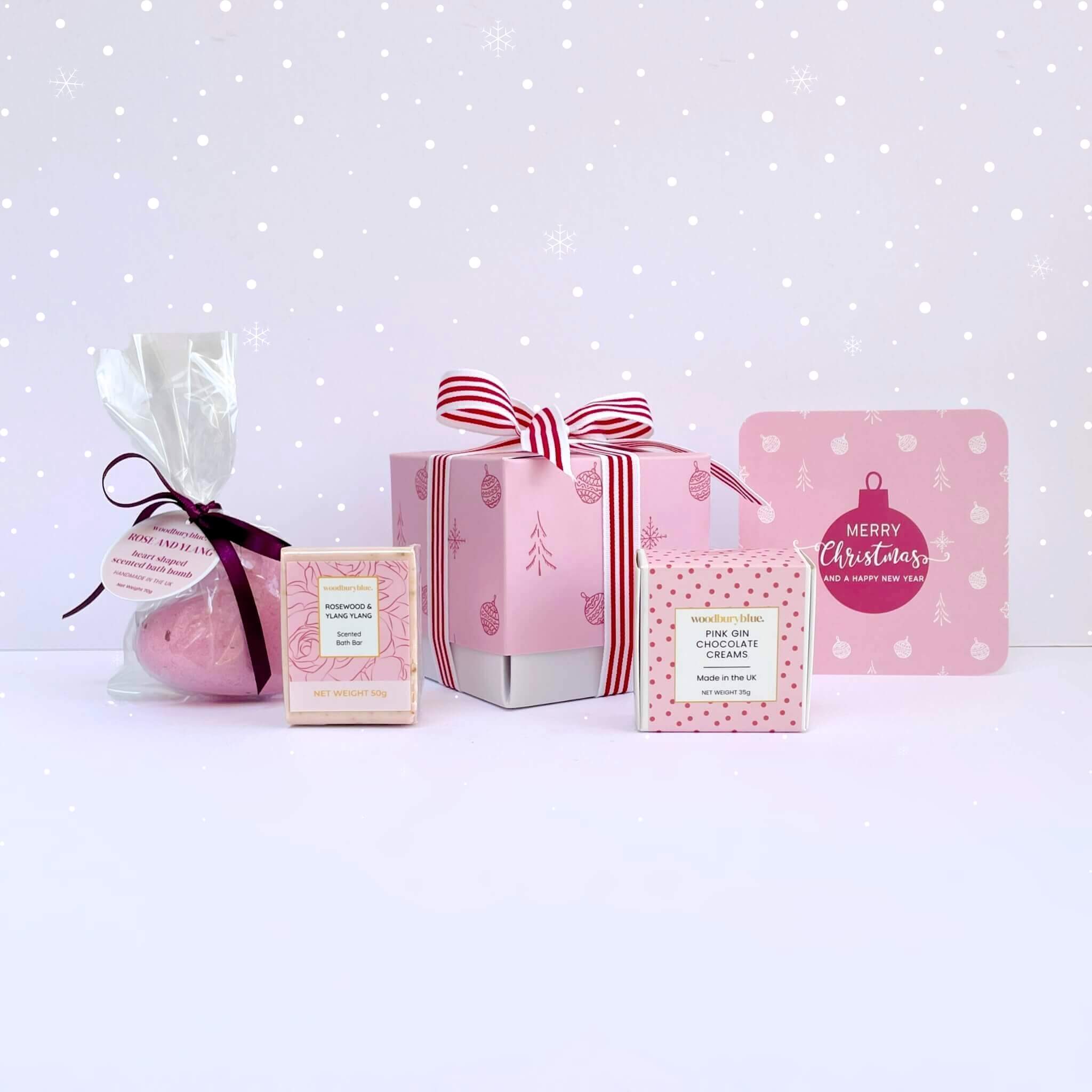 Rosy Glow Christmas Gift Box with pink bath bomb, rosewood soap, pink gin chocolate creams, pink ribbon gift box, and matching pink bauble Merry Christmas card on snowy background.