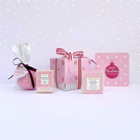 Rosy Glow Christmas Gift Box with pink bath bomb, rosewood soap, pink gin chocolate creams, pink ribbon gift box, and matching pink bauble Merry Christmas card on snowy background.