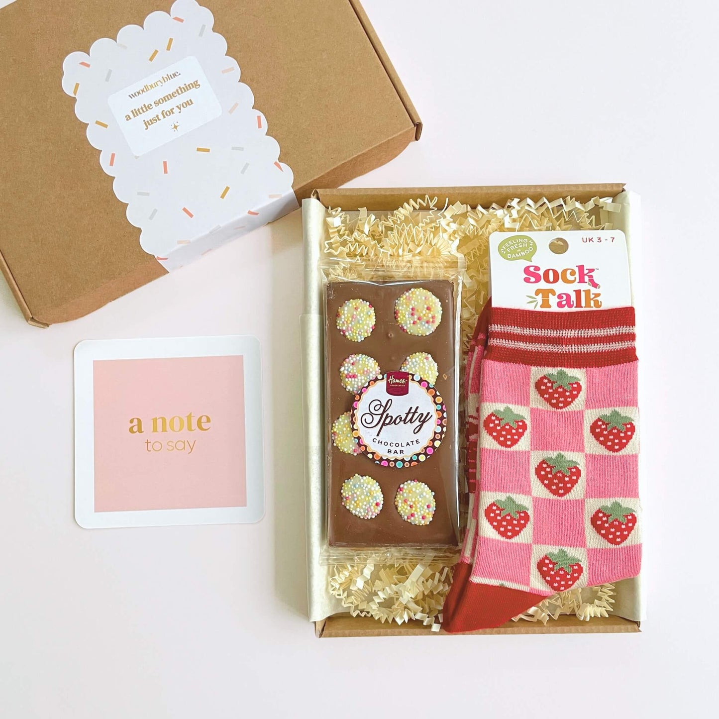Open letterbox gift box showing strawberry socks, spotty milk chocolate bar and a pink note card arranged inside shred packing.