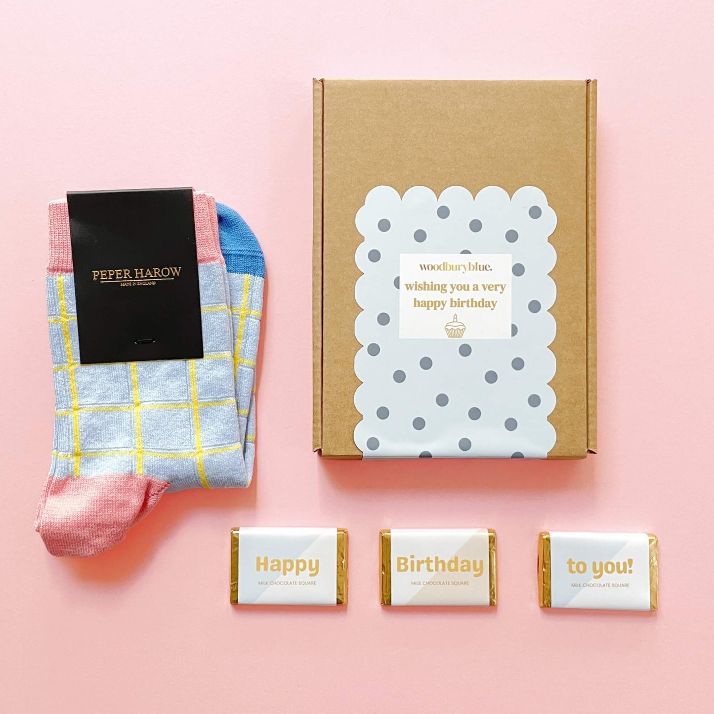 Flat lay of the Socks and Chocs Letterbox Gift – Birthday Wishes, showing blue grid Peper Harow socks, three Happy Birthday milk chocolate squares, and a polka dot birthday gift box on a pink background.
