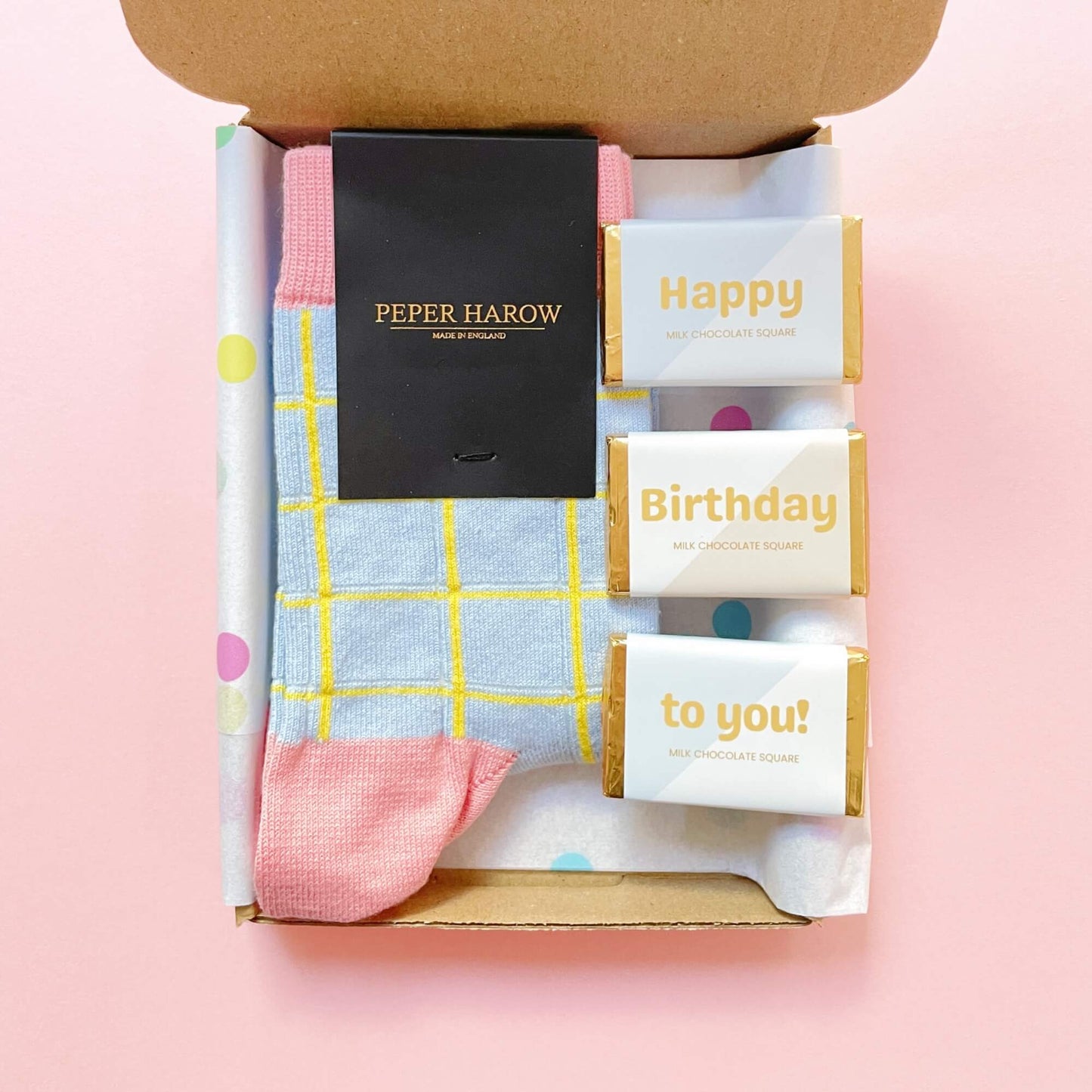 Close-up of the Socks and Chocs Letterbox Gift – Birthday Wishes, showing blue grid Peper Harow socks beside three Happy Birthday milk chocolate squares inside a brown gift box with polka dot tissue paper.