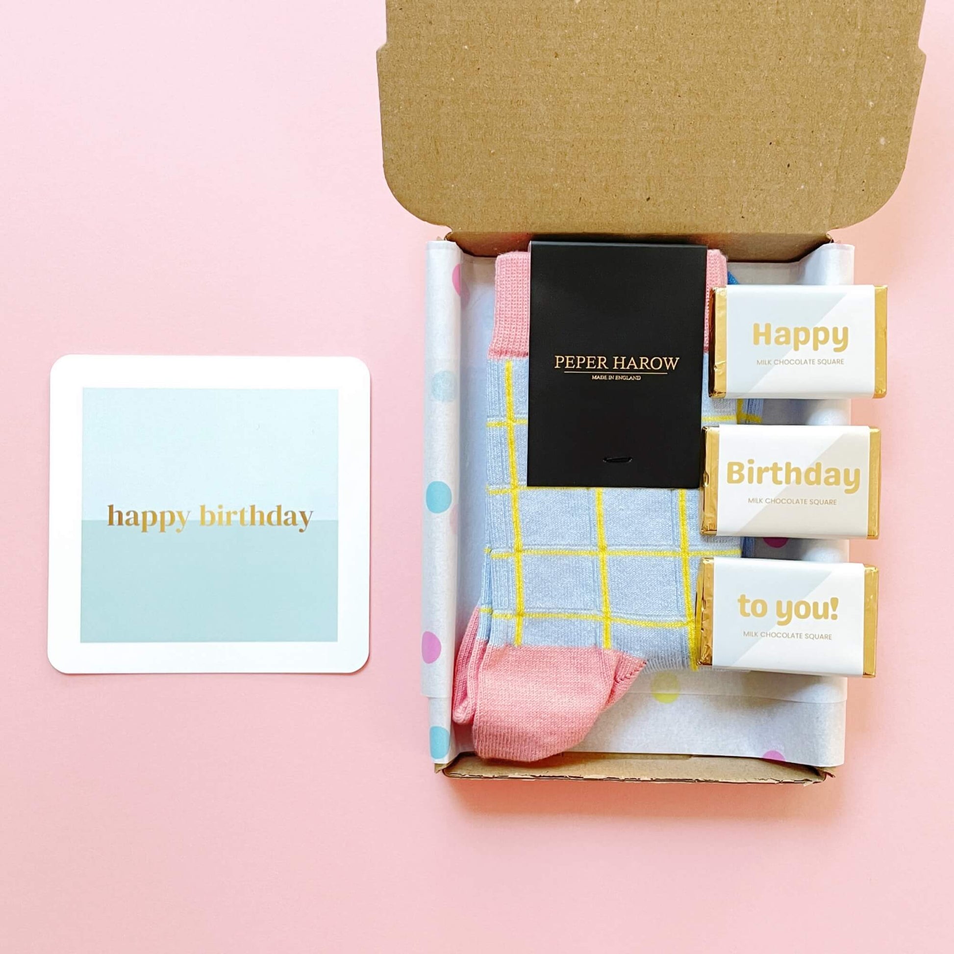 Open flat lay of the Socks and Chocs Letterbox Gift – Birthday Wishes, featuring blue grid Peper Harow socks, three Happy Birthday milk chocolate squares, and a mint and gold birthday card on a pink background.