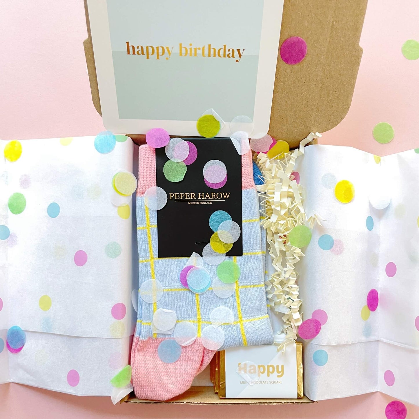 Open view of the Socks and Chocs Letterbox Gift – Birthday Wishes, showing Peper Harow blue grid socks, a Happy Birthday card, and milk chocolate squares surrounded by colourful confetti and tissue paper.