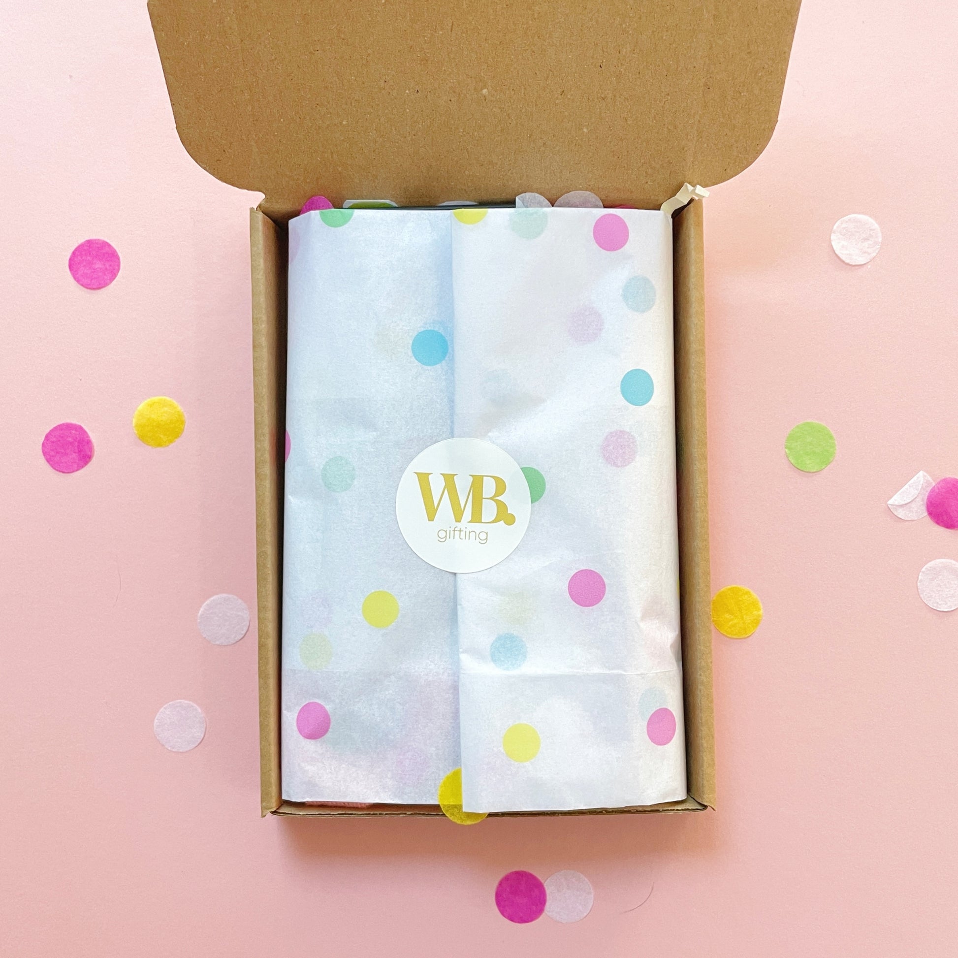 Birthday letterbox gift from Woodbury Blue wrapped in white polka dot tissue paper with a WB Gifting sticker, surrounded by colourful confetti on a pink background.