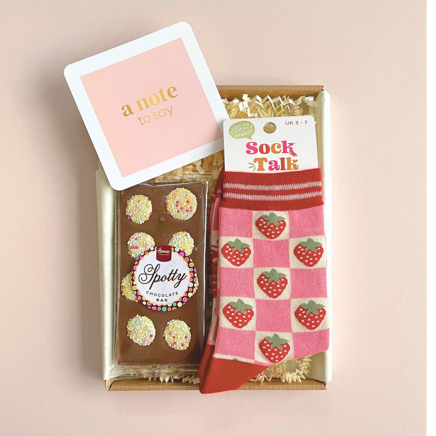 Strawberry Dreams gift box opened to show strawberry socks, sprinkle chocolate bar and a pink note card on soft pink background.