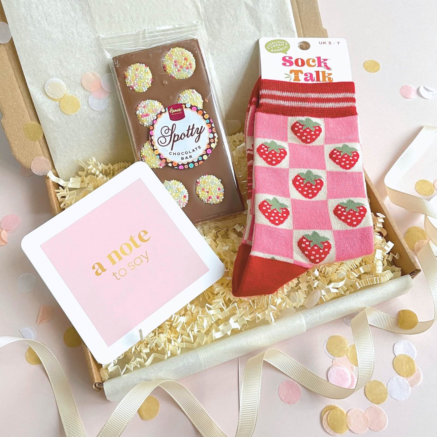 Open letterbox gift filled with strawberry socks and spotty chocolate bar, surrounded by pastel confetti and ribbon.