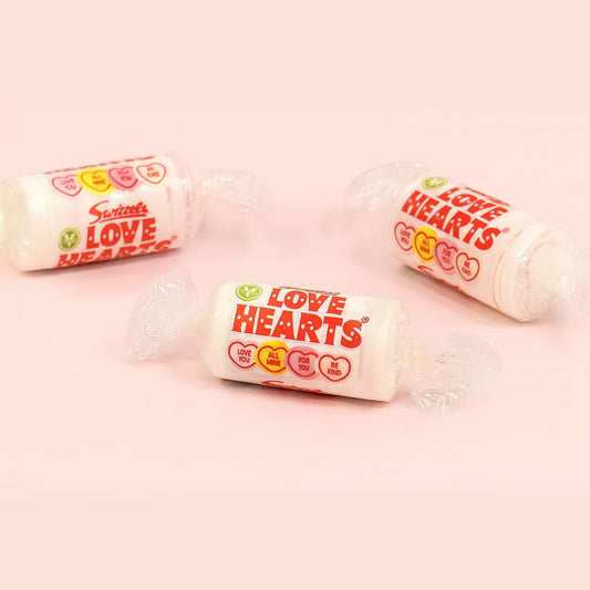 Three mini rolls of Swizzels Love Hearts sweets displayed on a soft blush pink background with clear wrappers and bold red logo