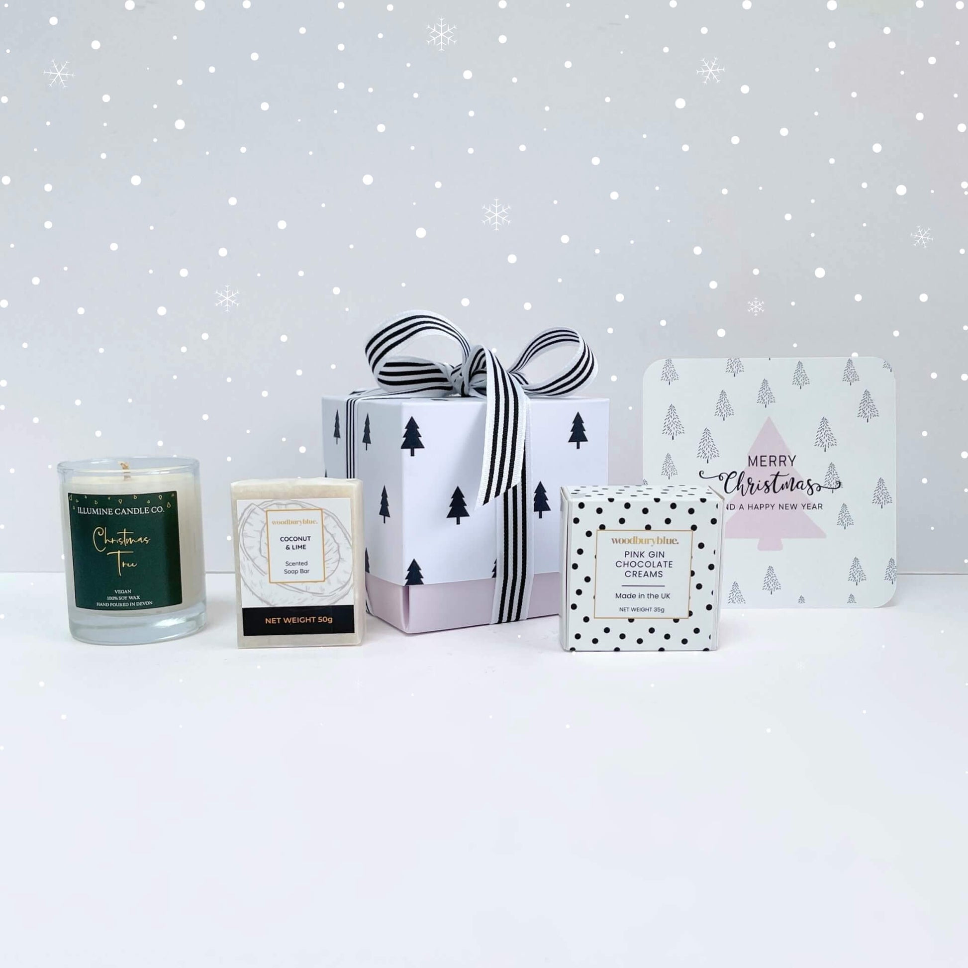 Warm Wishes Christmas Gift Box with coconut & lime soap, Christmas tree candle, pink gin chocolate creams and pink tree notecard in a black tree gift box on a snowy background.