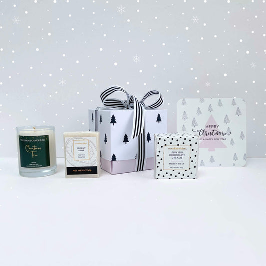Warm Wishes Christmas Gift Box with coconut & lime soap, Christmas tree candle, pink gin chocolate creams and pink tree notecard in a black tree gift box on a snowy background.