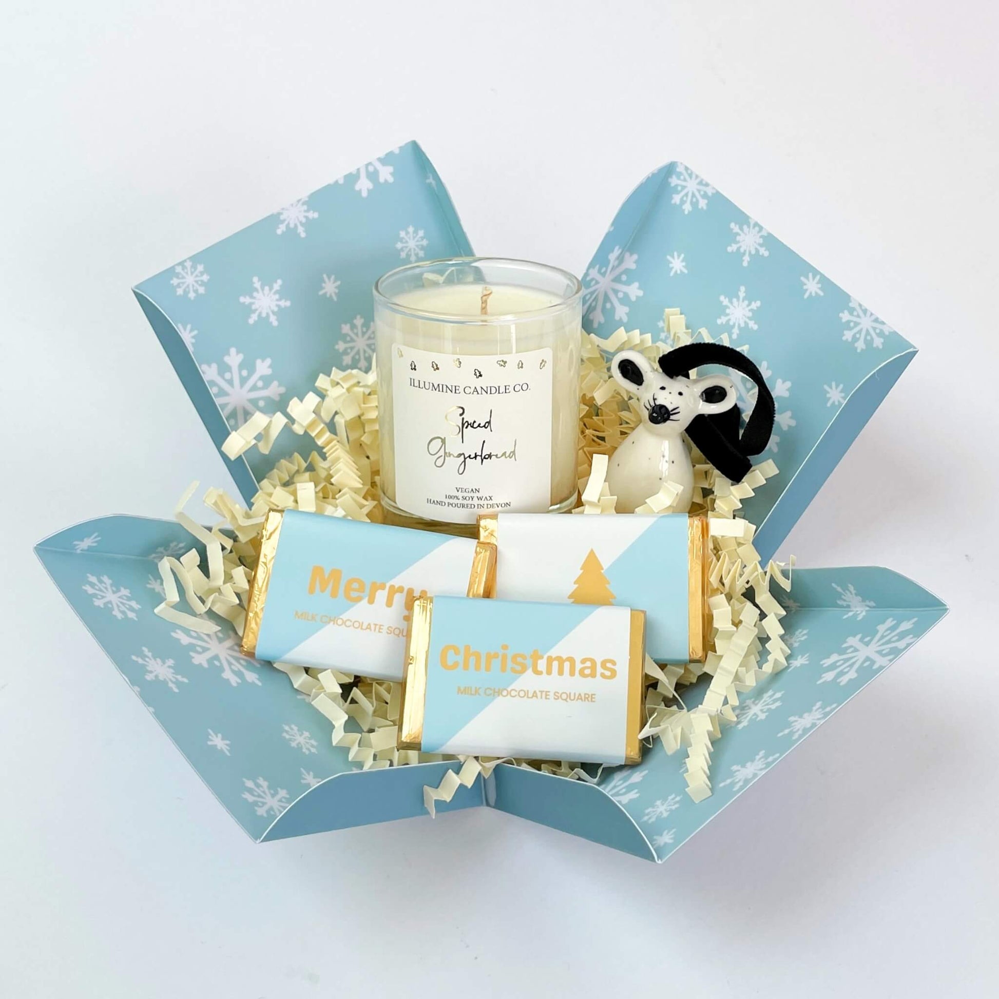 Winter Glow Christmas Gift Box shown open with shredded paper filling, a spiced gingerbread soy candle, ceramic mouse ornament, and three pale blue and gold chocolate squares inside a snowflake-print box.