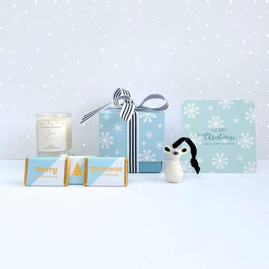 Winter Glow Christmas Gift Box styled with snowflake card, soy candle, blue gift box tied with striped ribbon, ceramic mouse ornament, and three mini milk chocolate squares on a snowy background.