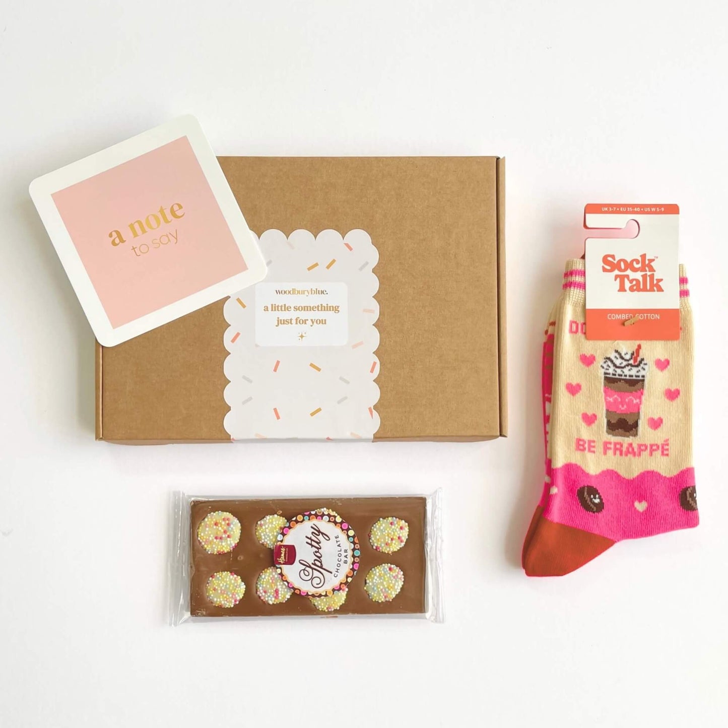 Flatlay of the Coffee Lovers letterbox gift showing a kraft box with a note card, Be Frappe socks and a Spotty chocolate bar laid out individually.