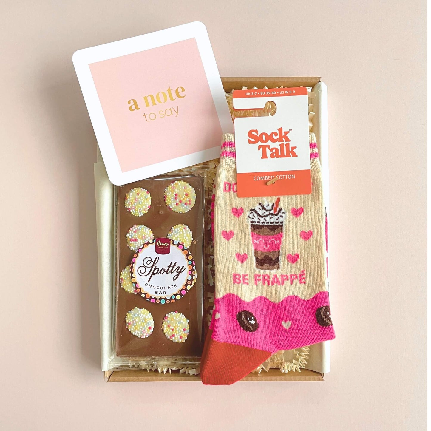 Top view of the Coffee Lovers Socks and Chocs gift with a Spotty chocolate bar and Be Frappe socks inside a kraft letterbox with pastel note card.