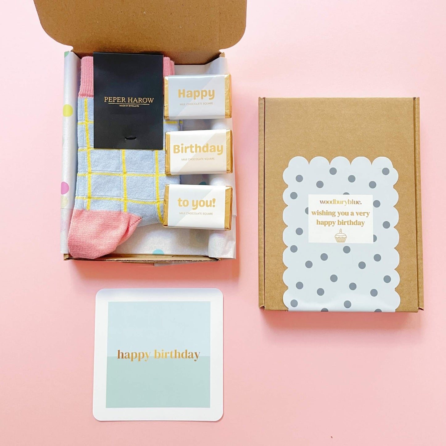Open birthday gift box showing Peper Harow socks, three happy birthday chocolates, and blue notecard on pink background