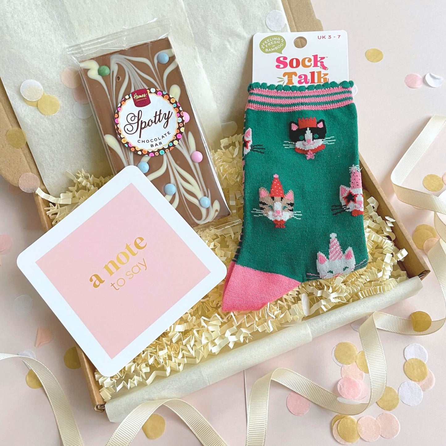 Lifestyle image of Party Cats Socks and Chocolates Letterbox Gift with pink notecard, chocolate bar and festive ribbon confetti styling
