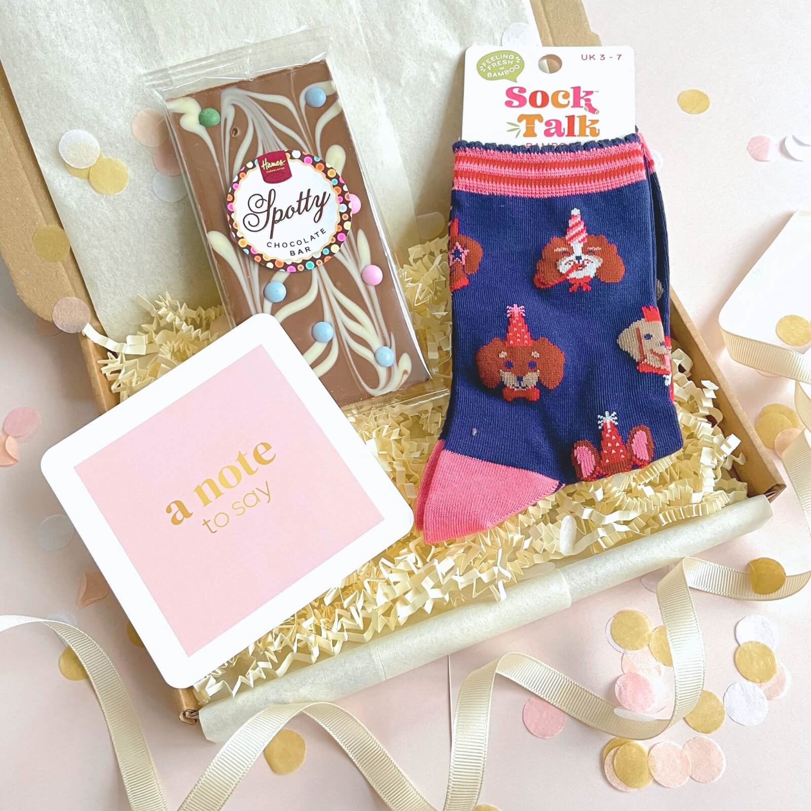 Lifestyle image of Party Dogs Socks and Chocolates Letterbox Gift with pink notecard, chocolate bar and confetti styling