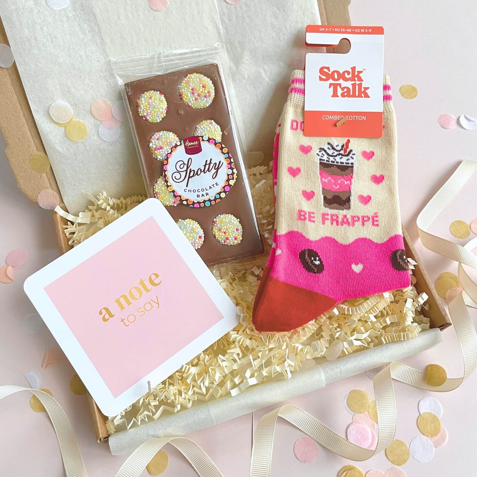 Coffee Lovers Socks and Chocs gift presented in an open kraft box with shred, Spotty chocolate bar, Be Frappe socks and a pastel pink note card surrounded by confetti.
