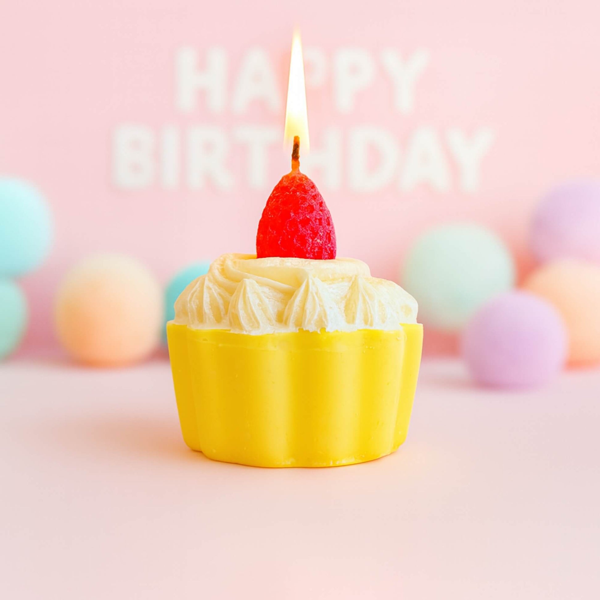 A strawberry cupcake candle with the wick lit, set against a pink birthday themed background with pastel pom poms behind it.