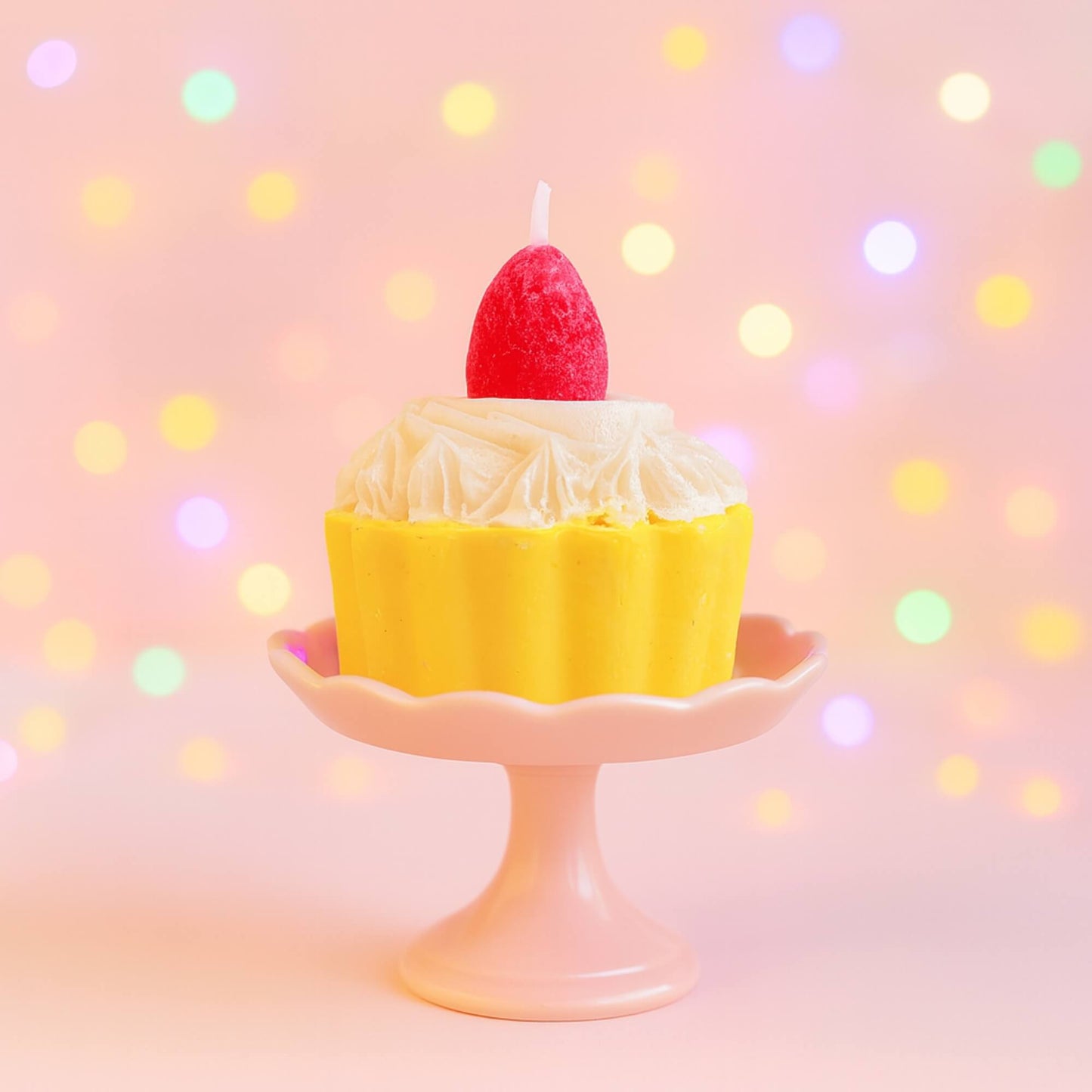 A strawberry cupcake candle placed on a small pink cupcake stand with colourful blurred lights in the background.