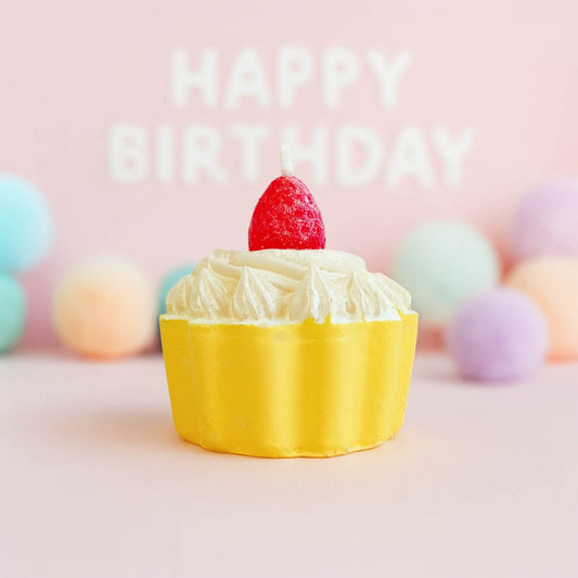 A yellow strawberry cupcake shaped candle with white frosting on a soft pink background and pastel decorations behind it.