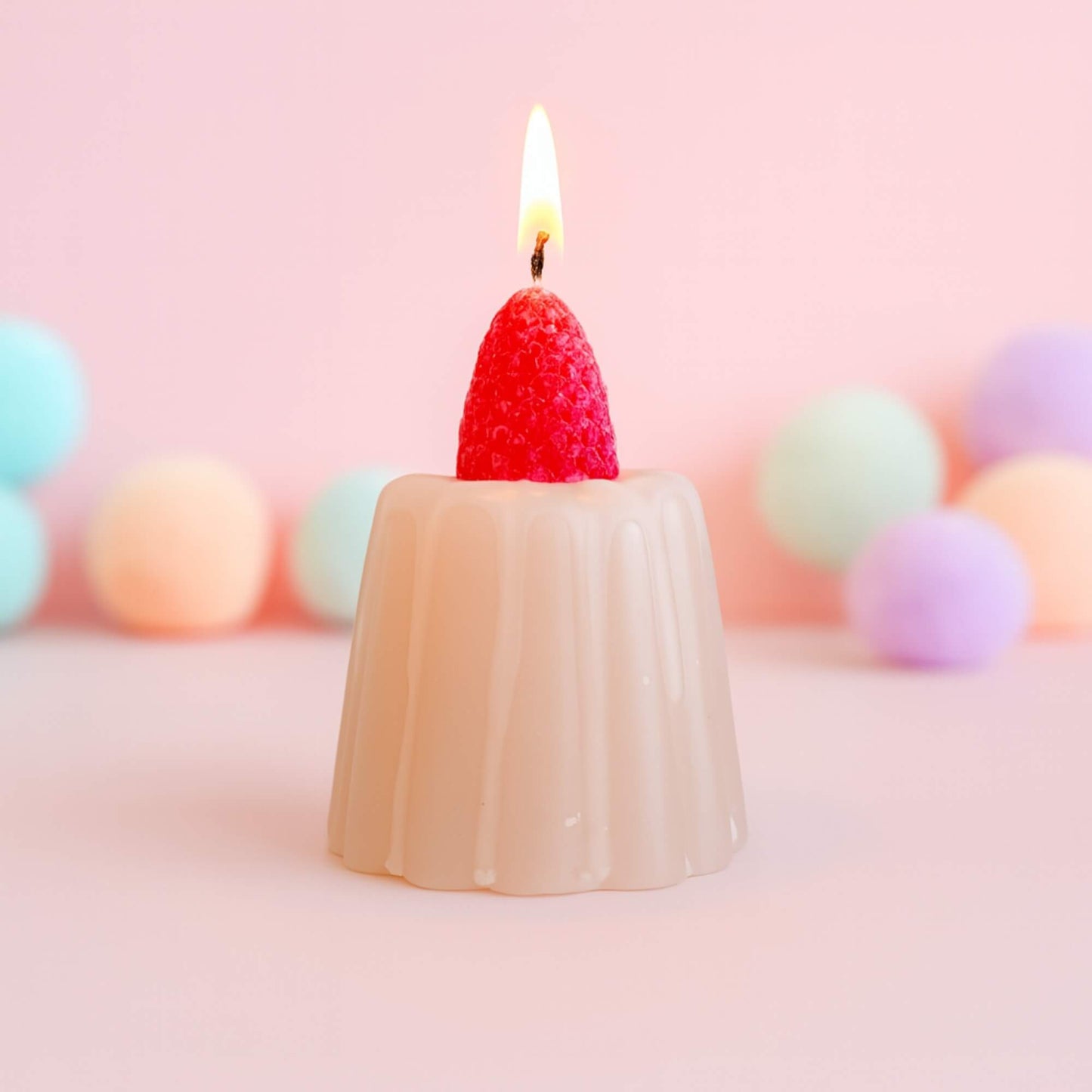 A strawberry jelly shaped candle with its wick lit, placed on a pink surface with pastel coloured pom poms in the background.