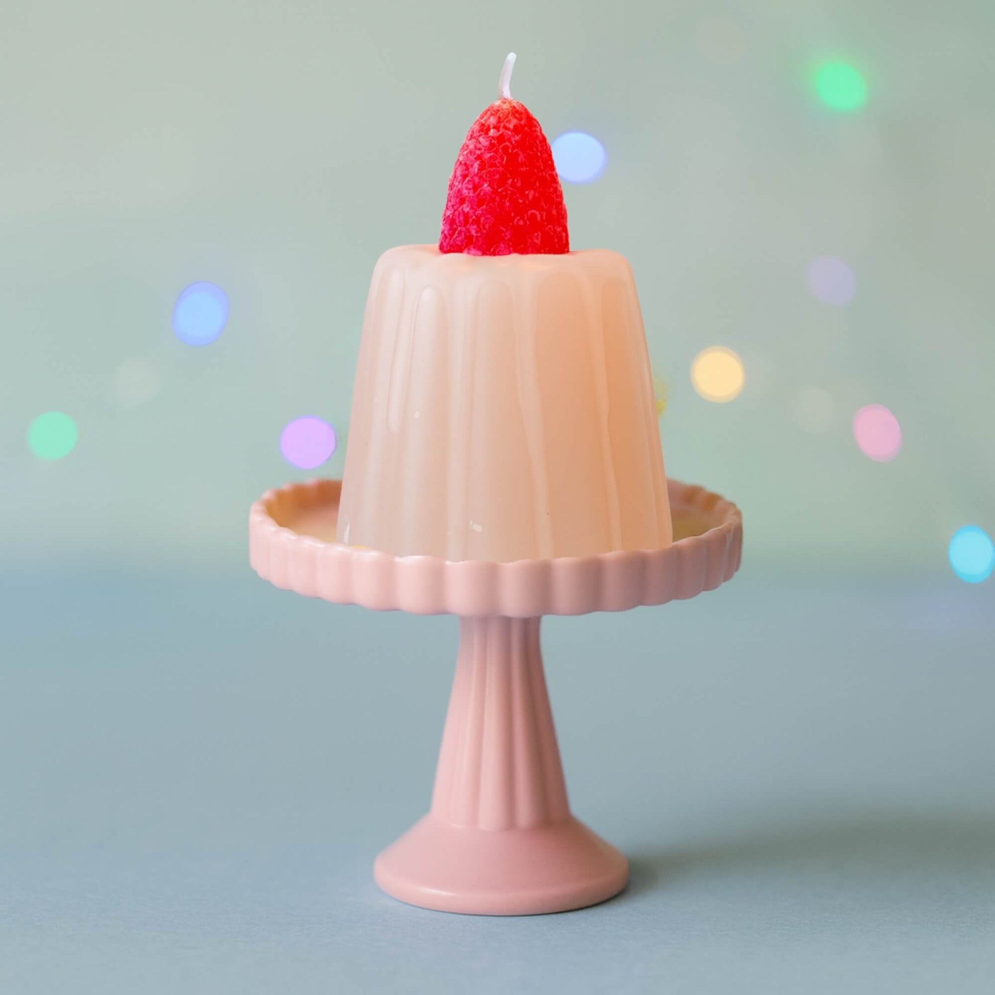 A strawberry jelly candle displayed on a small pink cupcake stand with a soft blue background and colourful blurred lights.