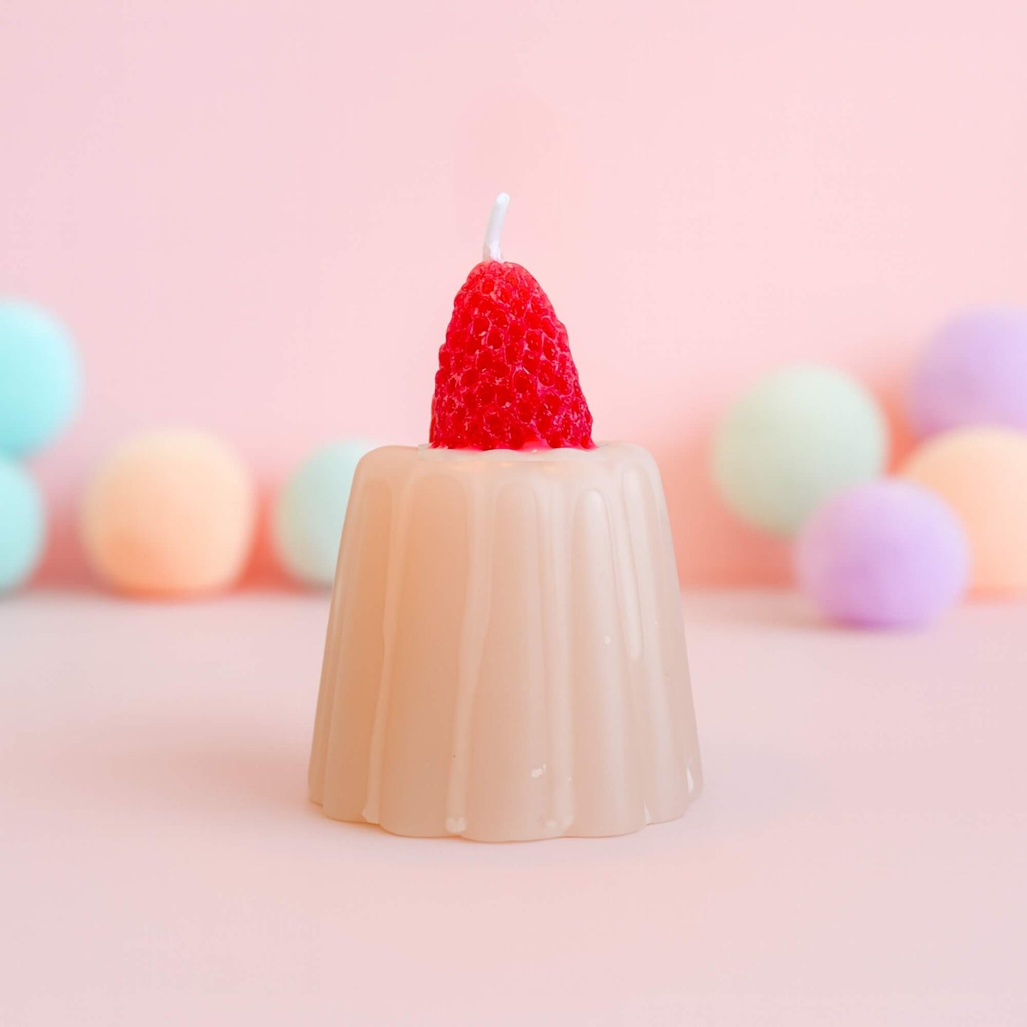 A strawberry jelly shaped candle with a bright red strawberry on top, set against a soft pink background with pastel spheres behind it.