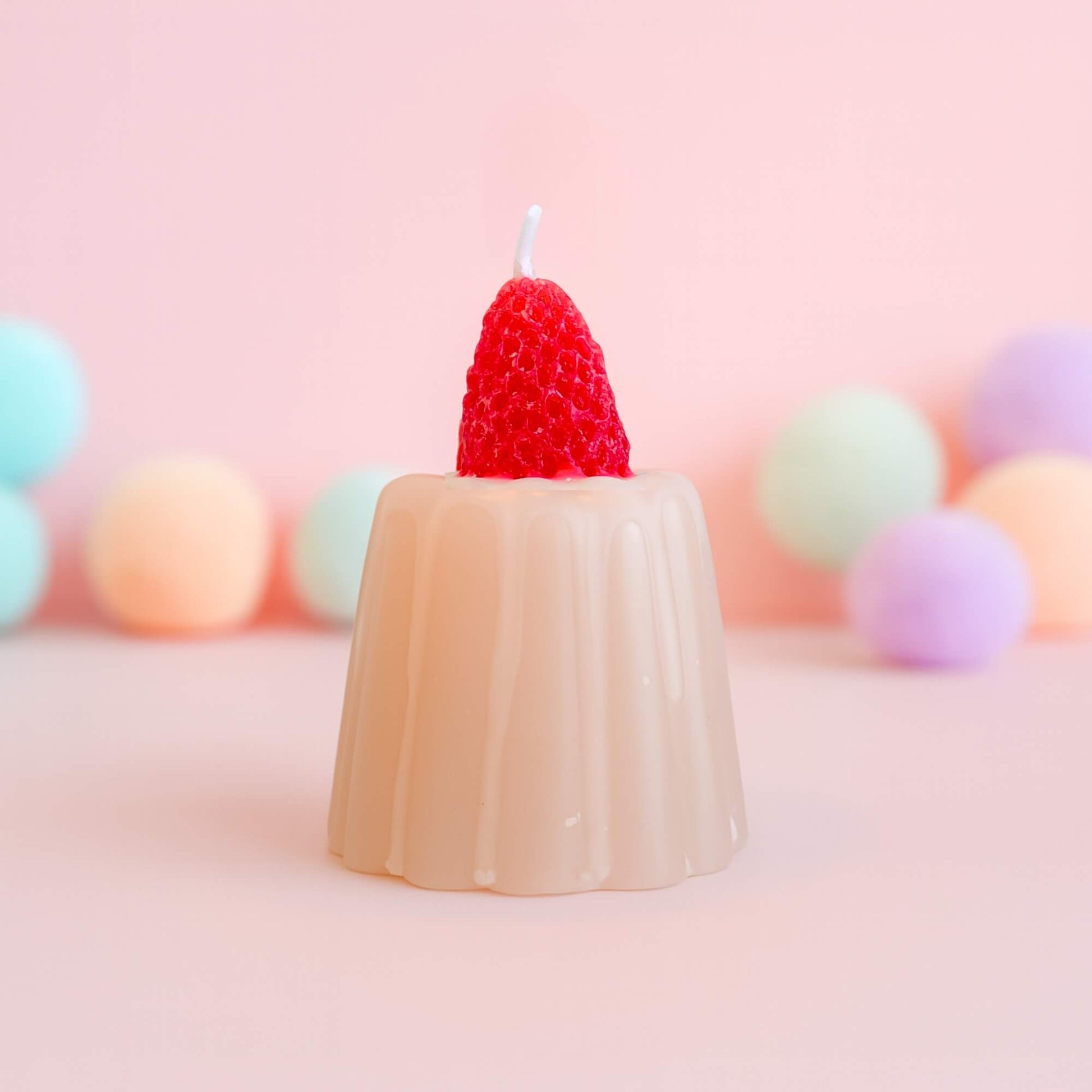 A strawberry jelly shaped candle with a bright red strawberry on top, set against a soft pink background with pastel spheres behind it.