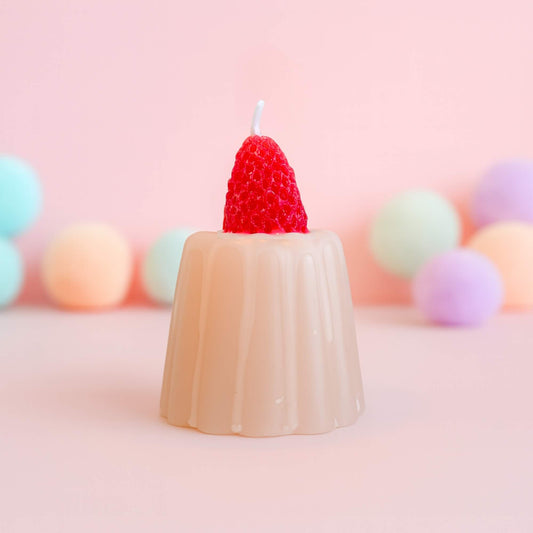 A strawberry jelly shaped candle with a bright red strawberry on top, set against a soft pink background with pastel spheres behind it.
