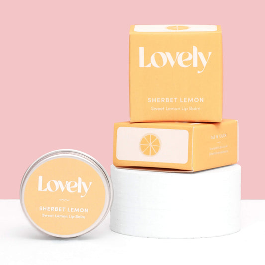 Lovely Skincare Sherbet Lemon Sweet Lemon Lip Balm tin and boxes stacked on white stand against pink background